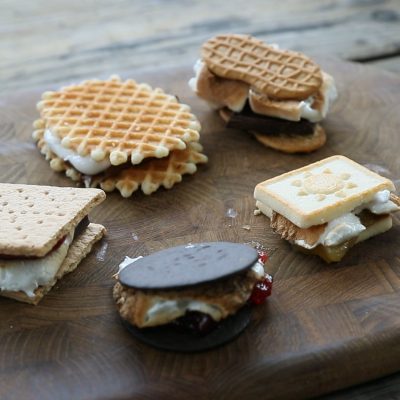A variety of gourmet s’mores made with different cookies and wafers, filled with melted marshmallows and chocolate, arranged on a rustic wooden board.