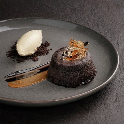 Elegant molten lava cake topped with edible gold leaf, served with vanilla ice cream and caramel sauce on a dark plate.
